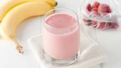 Strawberry smoothies in a clear glass sits on a white napkin with a bag of frozen strawberries and two bananas in the background
