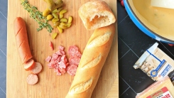 A pot of fondue next to chopped pickles, bread, sausage and herbs on a cutting board.