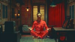 A still from THe Wonderful Story of Henry Sugar featuring Benedict Cumberbatch in an orange jumpsuit sitting crosslegged on an ornate rug in a room with dark paneled wood walls and fancy furnishings