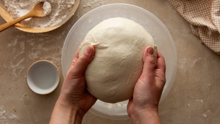 Hands holding a ball of bread dough over a bowl.