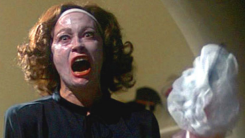 A screenshot of Faye Dunaway, face covered in cold cream and brandishing a wire hanger, from the film Mommie Dearest