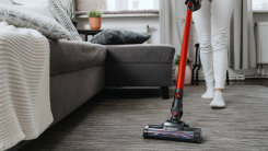 A person seen from the waist down vacuuming a carpet with a stick vacuum