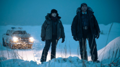 A screenshot from the True Detective season 4 trailer with Jodie Foster and Kali Reis in police uniforms and warm coats standing in a snowy field, illuminated by the beams of their police cruiser