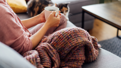 A woman sitting on a couch at home with a blanket, a hot drink, and a cute cat