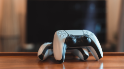 two playstation 5 controllers on a wooden table with a TV in the background