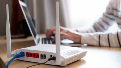 A close-up shot of a router. Behind, out of focus, is a person using their laptop