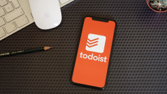 A phone displaying the Todoist logo, lying on an organized desk among computer accessories and a pencil.