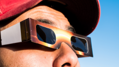 A close-up shot of a man outside who is wearing a red ball cap and a pair of solar eclipse glasses